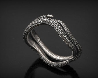 Puede incluir: Un anillo plateado con un diseño de tentáculo de pulpo. El anillo presenta texturas detalladas, incluyendo pequeños círculos en forma de ventosas y patrones acanalados. El anillo se encuentra sobre un fondo oscuro, resaltando su intrincado diseño.
