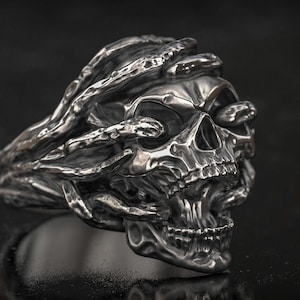 Könnte beinhalten: Ein detaillierter silberner Totenkopfring. Die Augenhöhlen des Schädels sind von einem rankenartigen Design bedeckt. Der Ring hat eine dunkle, strukturierte Oberfläche, die einen gotischen oder Biker-Look erzeugt.