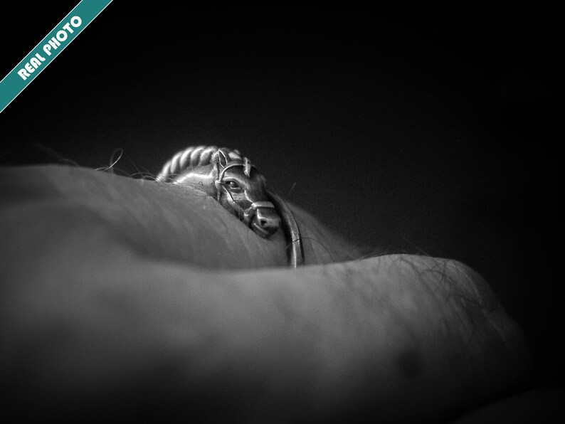 May include: A silver-toned ring featuring a horse head design. The horse head has detailed features, including a bridle. The ring is set against a dark background, creating a striking contrast. The image is labeled "REAL PHOTO".