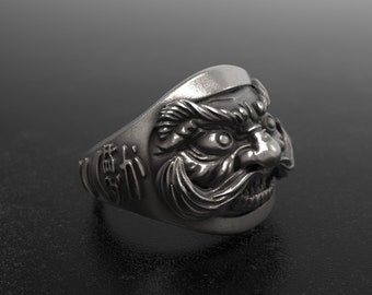 Könnte beinhalten: Ein silberfarbener Ring mit detailliertem Gesichtsdesign. Der Ring zeigt ein stilisiertes Gesicht mit markantem Schnurrbart und Bart. Der Ring hat japanische Schriftzeichen an der Seite. Der Ring ist vor einem dunklen Hintergrund platziert.