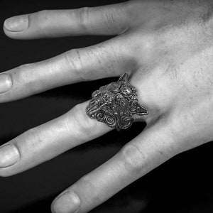 May include: A detailed silver ring featuring a wolf's head design. The ring is worn on a hand, showcasing the intricate details of the wolf's face and the ring's overall craftsmanship. The ring is a piece of jewelry.