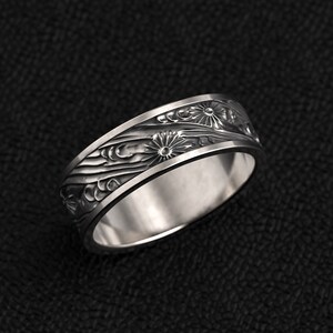 Könnte beinhalten: Ein silberner Ring mit floralem Design. Der Ring hat ein detailliertes Muster aus Blumen und fließenden Linien, die um das Band geschnitzt sind. Der Ring ist vor einem dunklen Hintergrund platziert, der die komplizierten Details des Schmucks hervorhebt.