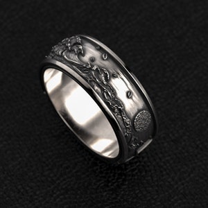 Könnte beinhalten: Ein silberner Ring mit detailliertem Reliefdesign. Der Ring zeigt ein Wellenmotiv mit einer Sonne und anderen Elementen. Der Ring wird auf einer schwarzen Oberfläche gezeigt.