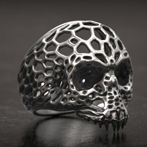 Könnte beinhalten: Ein silberner Totenkopfring mit einem einzigartigen, durchbrochenen Design. Der Ring zeigt eine detaillierte Totenkopfform mit einem wabenartigen Muster und scharfen Zähnen. Der Ring wird auf einer dunklen, strukturierten Oberfläche präsentiert.