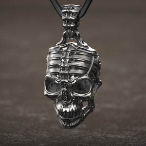 Puede incluir: Colgante de calavera plateada detallada con un cordón oscuro. El colgante presenta intrincados detalles esqueléticos, incluyendo la caja torácica y los rasgos faciales. El diseño de la calavera está muy estilizado, dándole una estética gótica única. El colgante está suspendido de un cordón negro.