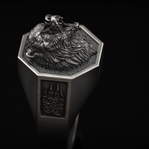 May include: A silver-colored ring featuring a detailed bear head design. The ring has an octagonal top with the bear's head and a rectangular side with a bear paw print. The ring is set against a black background.