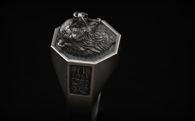 May include: A silver-colored ring featuring a detailed bear head design. The ring has an octagonal top with the bear's head and a rectangular side with a bear paw print. The ring is set against a black background.