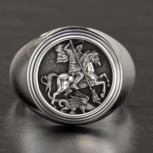Könnte beinhalten: Silberner Ring mit detailliertem Relief eines Ritters zu Pferd, der einen Drachen ersticht. Der Ring hat einen polierten, abgerundeten Ring und eine kreisförmige Oberfläche mit dem geprägten Design. Der Ritter ist in Rüstung dargestellt.