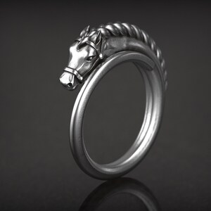 May include: A silver-colored ring featuring a horse head design. The horse head is detailed with a bridle and a braided mane. The ring curves around to form a complete circle. The ring is displayed against a dark background.