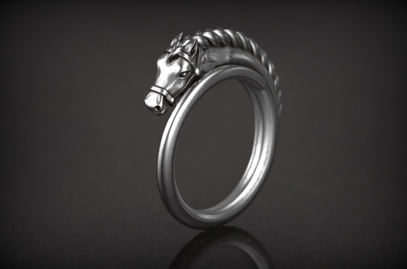 May include: A silver-colored ring featuring a horse head design. The horse head is detailed with a bridle and a braided mane. The ring curves around to form a complete circle. The ring is displayed against a dark background.