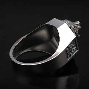 May include: A silver-colored ring with a detailed lion's head design on top. The ring has a rectangular face with an embossed emblem on the side. The ring is shown against a dark background.
