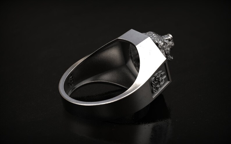 May include: A silver-colored ring with a detailed lion's head design on top. The ring has a rectangular face with an embossed emblem on the side. The ring is shown against a dark background.