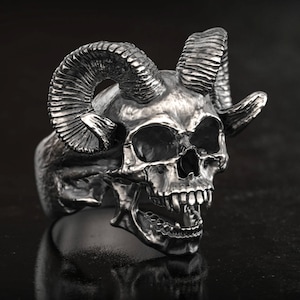 Könnte beinhalten: Ein silberfarbener Totenkopfring mit großen, gebogenen Hörnern. Der Ring weist detaillierte Gesichtszüge auf, darunter Augenhöhlen, eine Nasenhöhle und einen weit geöffneten Mund mit Zähnen. Der Ring ist vor einem dunklen Hintergrund platziert.