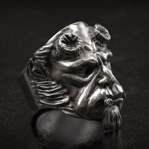 May include: A silver-toned ring featuring a detailed, sculpted face with horns, a goatee, and a cigar. The ring has a dark, textured background, highlighting the intricate design and metallic sheen of the jewelry.