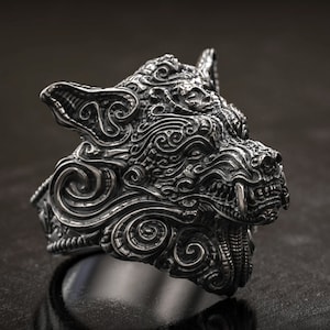 May include: A detailed silver ring shaped like a wolf's head. The ring features intricate carvings and a menacing expression, with visible teeth and pointed ears. The design is highly textured and detailed.