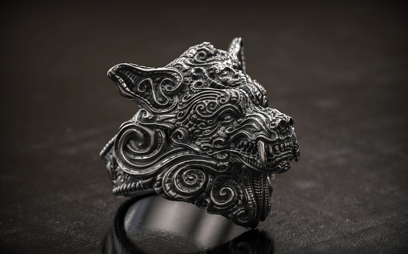 May include: A detailed silver ring shaped like a wolf's head. The ring features intricate carvings and a menacing expression, with visible teeth and pointed ears. The design is highly textured and detailed.