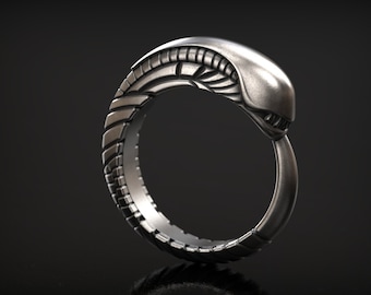 May include: A silver-colored ring with a detailed design resembling an alien creature. The ring features a segmented band and a head-like structure at the top, with intricate detailing. The ring is displayed against a dark background.