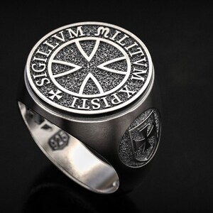 Könnte beinhalten: Ein silberner Ring mit detailliertem Design. Der Ring hat ein rundes Emblem mit dem Text "SIGILLVM MILITVM XPISTI" und einem Kreuzsymbol. Der Ring ist vor einem dunklen Hintergrund platziert, der seine filigranen Details hervorhebt.