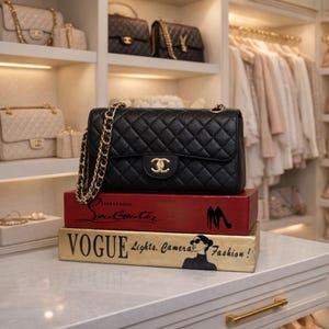 May include: A black quilted leather handbag with a gold chain strap, displayed on decorative books. The books are red and gold, with the words "Christian Louboutin" and "VOGUE Lights, Camera, Fashion!" printed on them. Other handbags are visible in the background.
