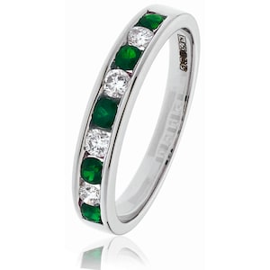 May include: A silver band ring with alternating emerald green and clear gemstones. The ring features a channel setting, with the gemstones set flush within the band. The ring is highly polished and reflects light.