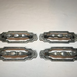 May include: Four antique silver drawer pulls with an ornate design. Each pull has a rectangular shape with a raised center section and a decorative handle.