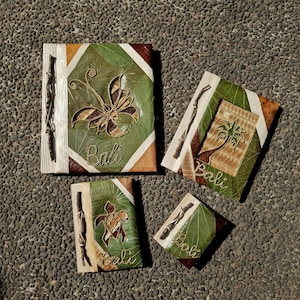Puede incluir: Cuatro diarios rectangulares con cubiertas de textura de hoja verde, cada uno con encuadernación de ramitas y la palabra Bali bordada en hilo blanco. Las cubiertas muestran diseños cosidos de una mariposa, una palmera y una tortuga.