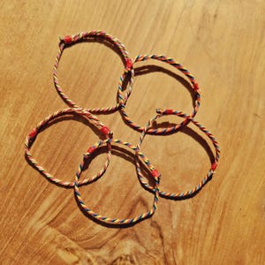 May include: Five thin bracelets made of twisted multi-colored cord in shades of red, yellow, blue, and white. Each circular bracelet features a small, solid red knot detail that secures the cord, creating a simple and lightweight design.