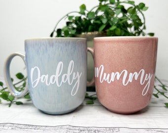 Könnte beinhalten: Zwei Keramikbecher mit einem Verlaufsglasur. Der Becher links ist hellblau mit dem Wort "Daddy" in weißen Buchstaben. Der Becher rechts ist rosa mit dem Wort "Mummy" in weißen Buchstaben.