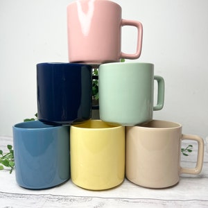 May include: Six ceramic mugs in different colours stacked in a pyramid shape. The mugs are light pink, navy blue, light green, light blue, yellow, and beige.