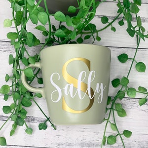 May include: A sage green ceramic mug with a gold letter "S" and the name "Sally" in white script. The mug has a curved handle and is surrounded by green leafy vines.