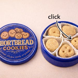 May include: A blue tin of shortbread biscuits, lid off, showing five biscuits in separate compartments. The lid is printed with "Shortbread Cookies" and smiling cookie illustrations. The text "Sweet clicks, zero crumbs" is also on the lid.