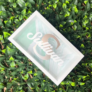 May include: A white framed shadow box displaying the name "Sullivan" in gold script. The background has a light green and white marbled pattern. The box is set against a backdrop of green foliage.