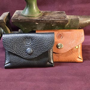 May include: Two small leather pouches, one black and one tan, with flap closures and metal snap buttons. Each pouch has a decorative emblem. The pouches are displayed in front of a metal anvil.