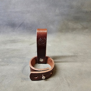 May include: A brown leather belt loop with a metal snap closure. The loop is designed to hold a belt or strap.