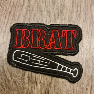 Pode incluir: Patch preto com a palavra "BRAT" em letras vermelhas e um desenho de taco de beisebol branco. O patch tem uma borda preta e é destinado a roupas ou acessórios.
