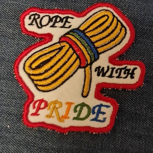 May include: Embroidered patch with a yellow rope design and a rainbow-coloured band. The patch reads "ROPE WITH PRIDE" in black and rainbow lettering, outlined in red and white.