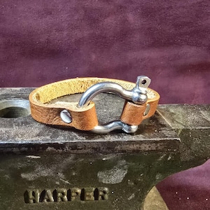 May include: A brown leather bracelet with a silver shackle clasp. The bracelet is made of light brown leather and features a sturdy, curved metal clasp. The bracelet is secured with two silver rivets. The bracelet is shown on a metal surface.