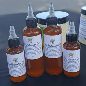 May include: Four bottles of Hair Restoration Oil with white labels and black caps. The bottles are filled with a brown liquid. The labels list ingredients like stinging nettle root and essential oils. A jar is visible in the background.