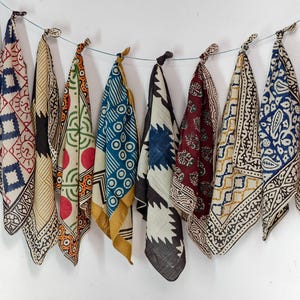 May include: A collection of ten square cotton bandanas hanging from a thin wire. Each bandana features a unique block-printed pattern in various colours including blue, red, yellow, black, and cream, showcasing geometric, floral, and abstract motifs.