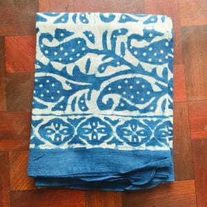 May include: Folded blue and white patterned textile. The fabric features a repeating design of stylized shapes and dots in white against a blue background. The bottom edge has a solid blue border. The textile is folded neatly.