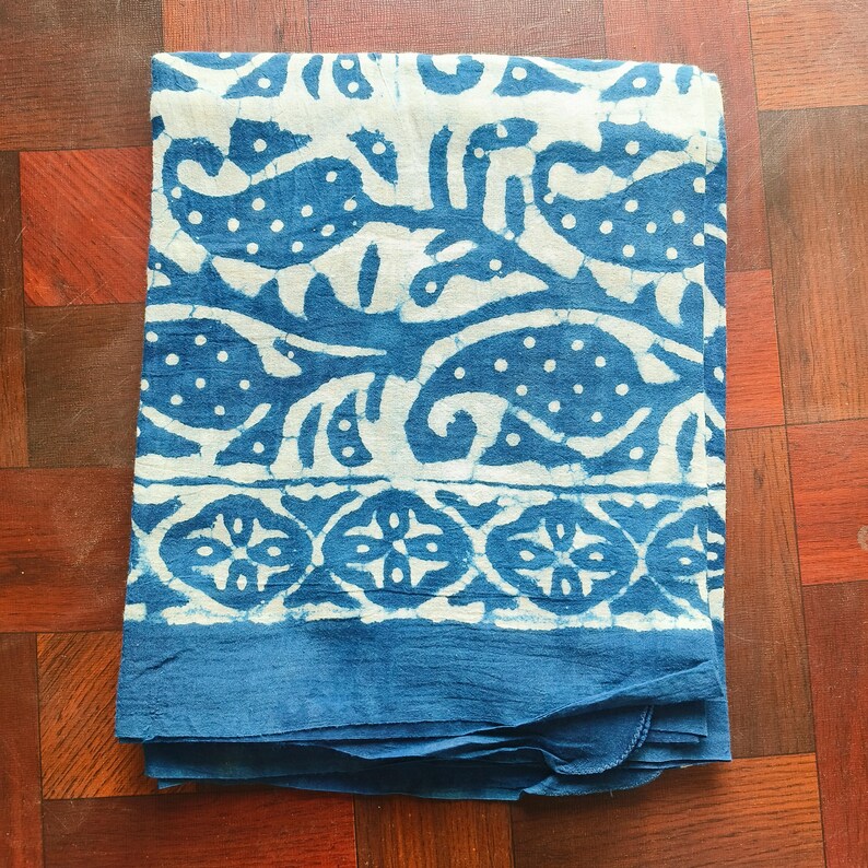 May include: Folded blue and white patterned textile. The fabric features a repeating design of stylized shapes and dots in white against a blue background. The bottom edge has a solid blue border. The textile is folded neatly.