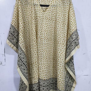 May include: A cream-colored kaftan with a V-neck and short sleeves. The fabric features a repeating pattern of small gold flowers and decorative black and white borders. The kaftan is hanging on a black hanger.