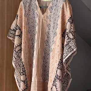 Op de afbeelding: Een perzikkleurige, bedrukte kaftan met een V-hals. De kaftan heeft een herhalend cirkelvormig en geometrisch ontwerp in marineblauw en zwart. De mouwen en randen zijn afgewerkt met een zwart-wit paisley patroon. Het kledingstuk hangt aan een zwarte hanger.