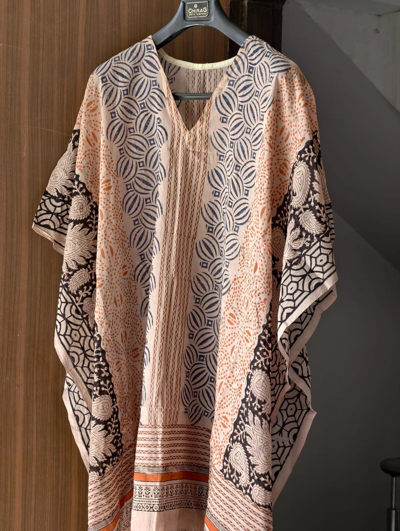 Op de afbeelding: Een perzikkleurige, bedrukte kaftan met een V-hals. De kaftan heeft een herhalend cirkelvormig en geometrisch ontwerp in marineblauw en zwart. De mouwen en randen zijn afgewerkt met een zwart-wit paisley patroon. Het kledingstuk hangt aan een zwarte hanger.