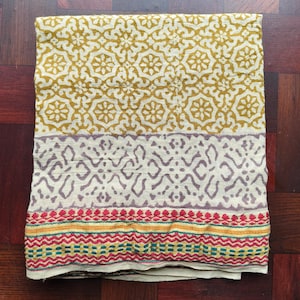 May include: Folded textile featuring a yellow floral pattern, a cream section with a purple geometric design, and a colourful embroidered border with red, yellow, and green threads. The fabric is neatly folded.