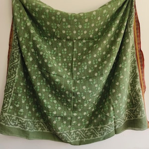 May include: A green textile with a floral pattern and decorative border. The fabric has a stitched appearance and a contrasting red and yellow border on one side. The textile is draped, showing its full design.