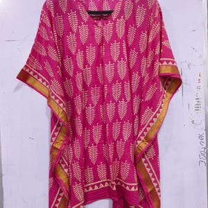 May include: A pink kaftan with a beige leaf pattern and gold trim. The lightweight fabric drapes loosely, with wide sleeves and a V-neck. The gold trim features a geometric design along the edges.