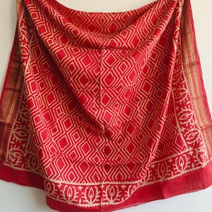 May include: A red scarf featuring a repeating diamond pattern in cream. The edges are adorned with a cream border showcasing floral and leaf designs. The fabric is draped, highlighting its texture. A colourful accessory.