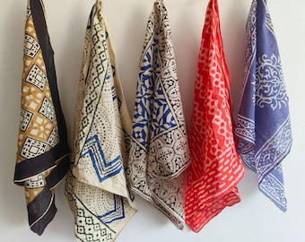 May include: Five patterned scarves in various colors hang against a white background. The scarves feature geometric and floral designs in shades of gold, black, blue, red, and purple. The fabric appears lightweight and flowing.