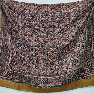 Puede incluir: Textil rectangular con un detallado estampado floral en rojo, azul y blanco sobre un fondo oscuro. El borde es de color amarillo mostaza liso. La tela está drapeada, mostrando su textura y diseño.